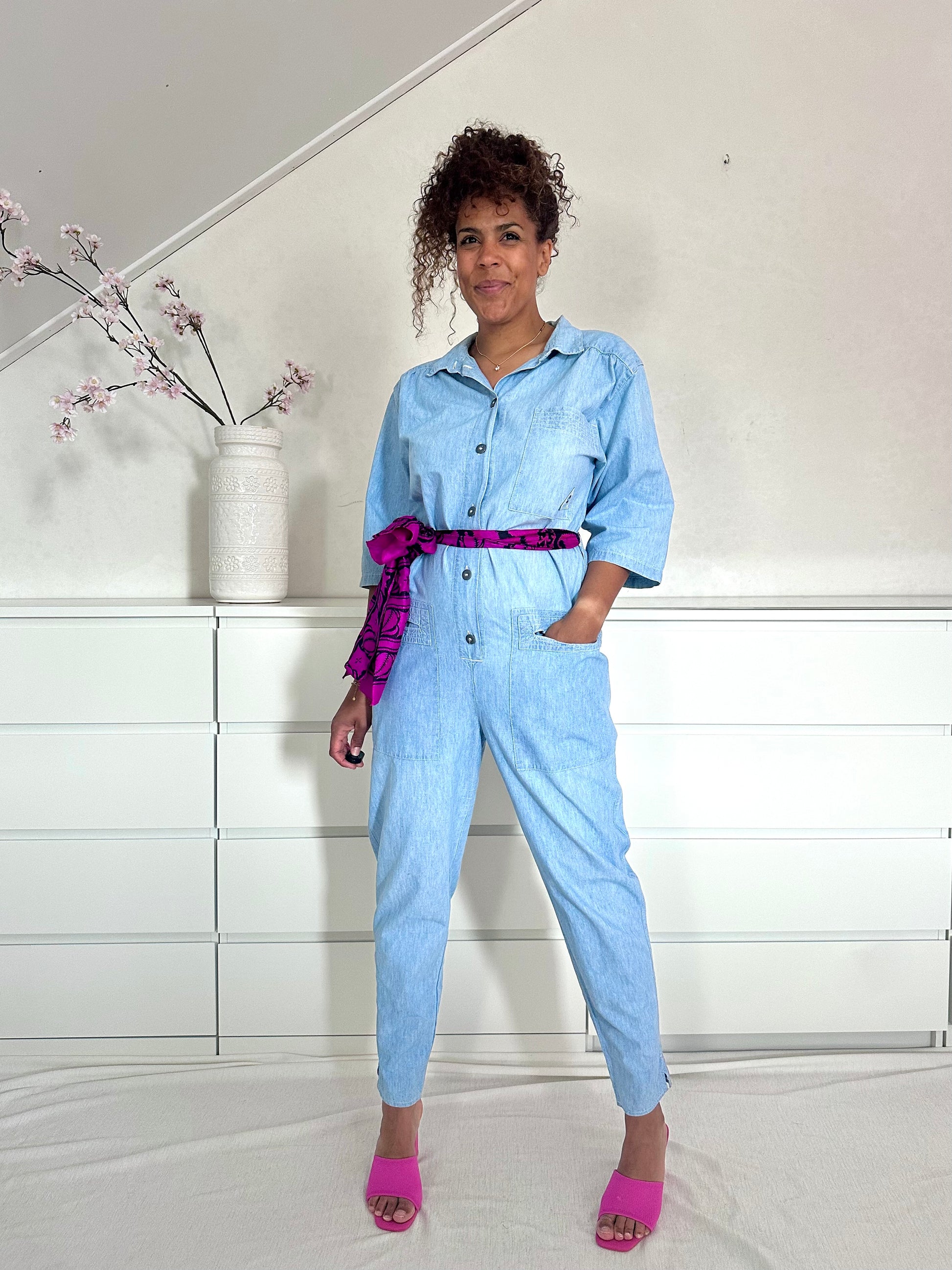 Vintage lichtblauwe spijkerjumpsuit met knopen en borstzakken, gestyled met een opvallende roze ceintuur.